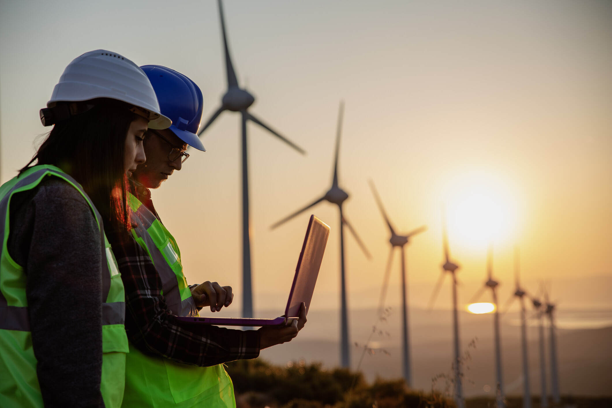 Young electrical engineer woman and business man standing in front of wind turbines checking and working about technical problems and writes the results of measurements with laptop pc in wind power plant electric energy station.